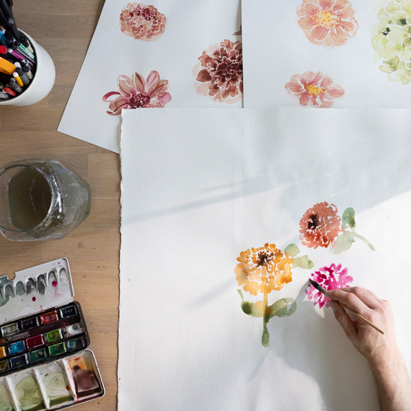 Person painting flowers on paper with watercolors on a wooden table.