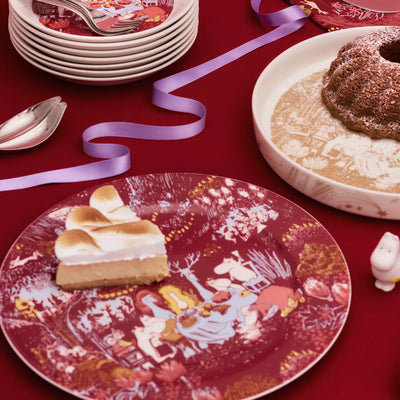 Decorative plate with dessert on a red tablecloth, surrounded by other plates and a cake.