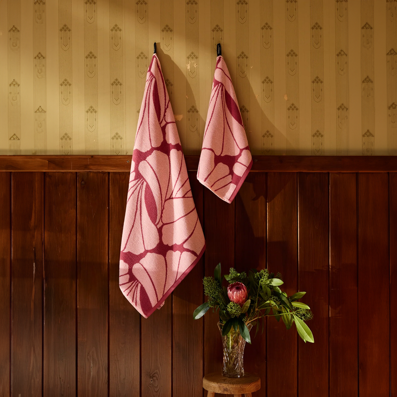 Two pink patterned towels hanging on a wooden wall with a vase of flowers below.