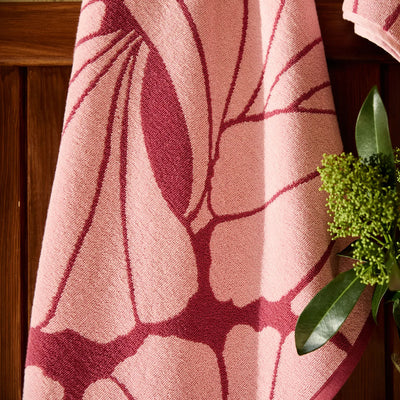 Pink towel with leaf pattern hanging on a wooden surface next to green leaves.