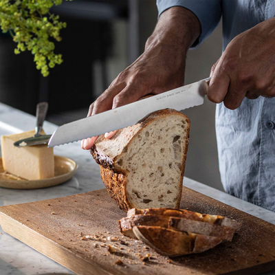 Person slicing bread on a wooden cutting board with a knife, with a plant and cheese block in the background.