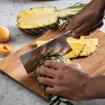 Person cutting a pineapple on a wooden cutting board with a knife.