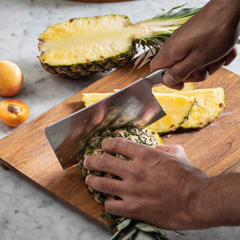 Person cutting a pineapple on a wooden cutting board with a knife.