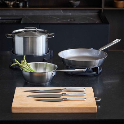 Set of stainless steel cookware including pots, pans, and a cutting board with knives on a dark surface.