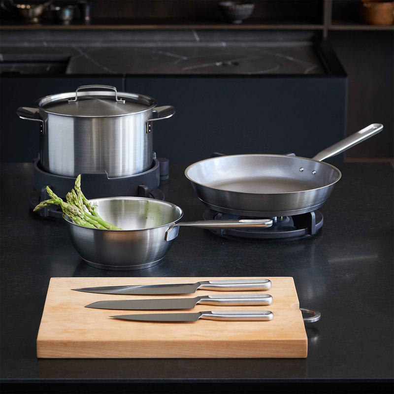 Set of stainless steel cookware including pots, pans, and a cutting board with knives on a dark surface.