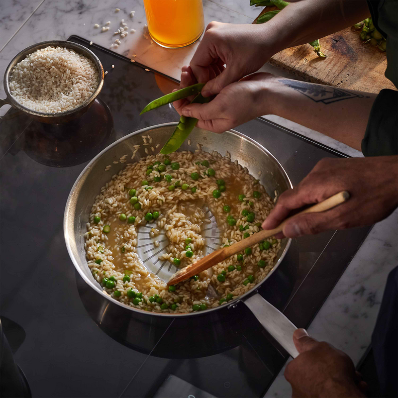Person cooking risotto with peas in a pan on a stove, with a glass of beer in the background.