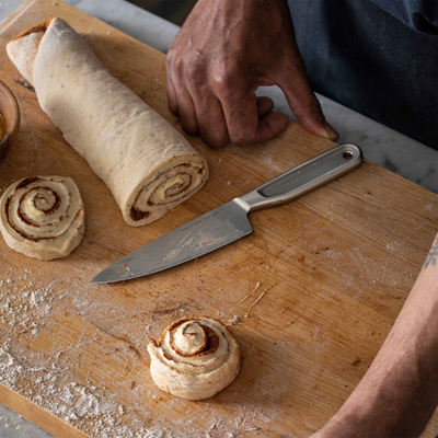Person cutting cinnamon rolls on a wooden board with a knife.