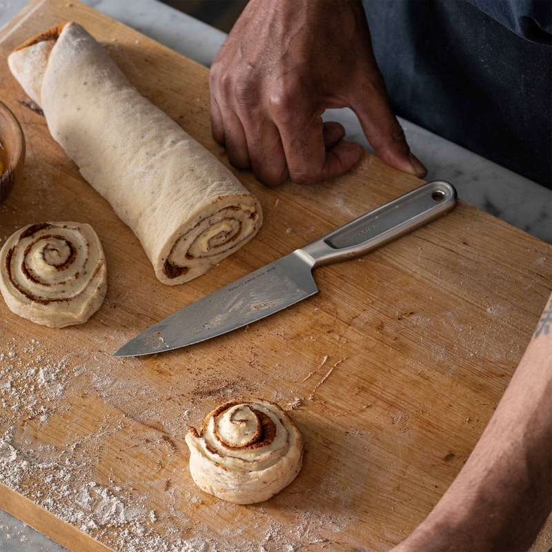 Person cutting cinnamon rolls on a wooden board with a knife.
