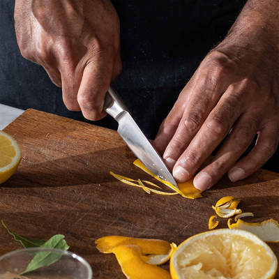 Person peeling a lemon on a wooden cutting board with a knife.
