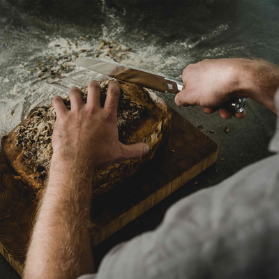 Person slicing bread on a wooden cutting board with a knife.