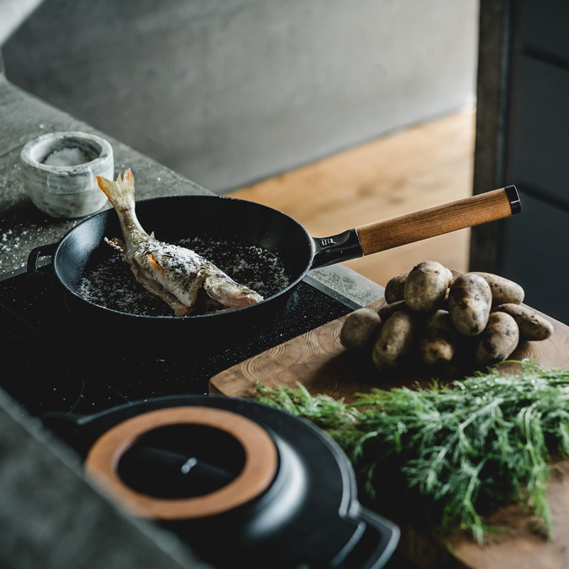 Frying pan with fish on a stove, surrounded by potatoes and herbs on a wooden surface.