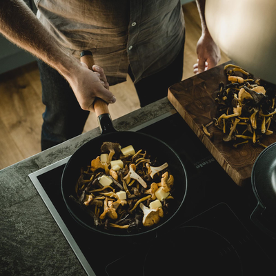 Person cooking mushrooms in a pan on a stove with a wooden cutting board nearby.