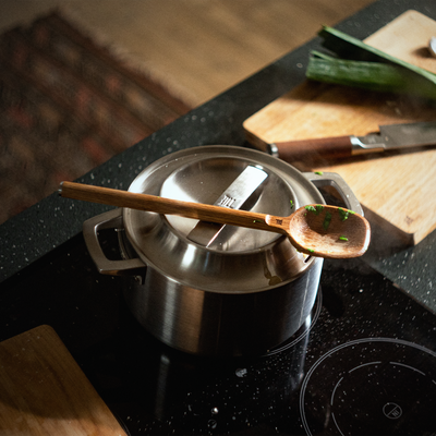 Stainless steel pot on a stovetop with a wooden spoon inside, surrounded by kitchen items.