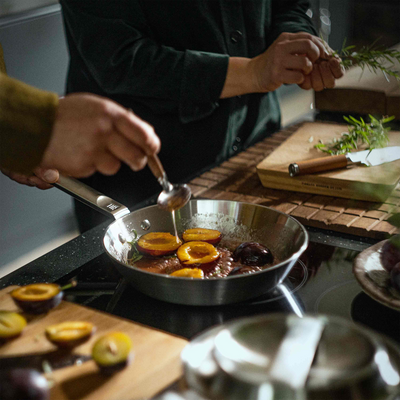 Frying pan with food on a stove, surrounded by ingredients and utensils on a wooden surface.