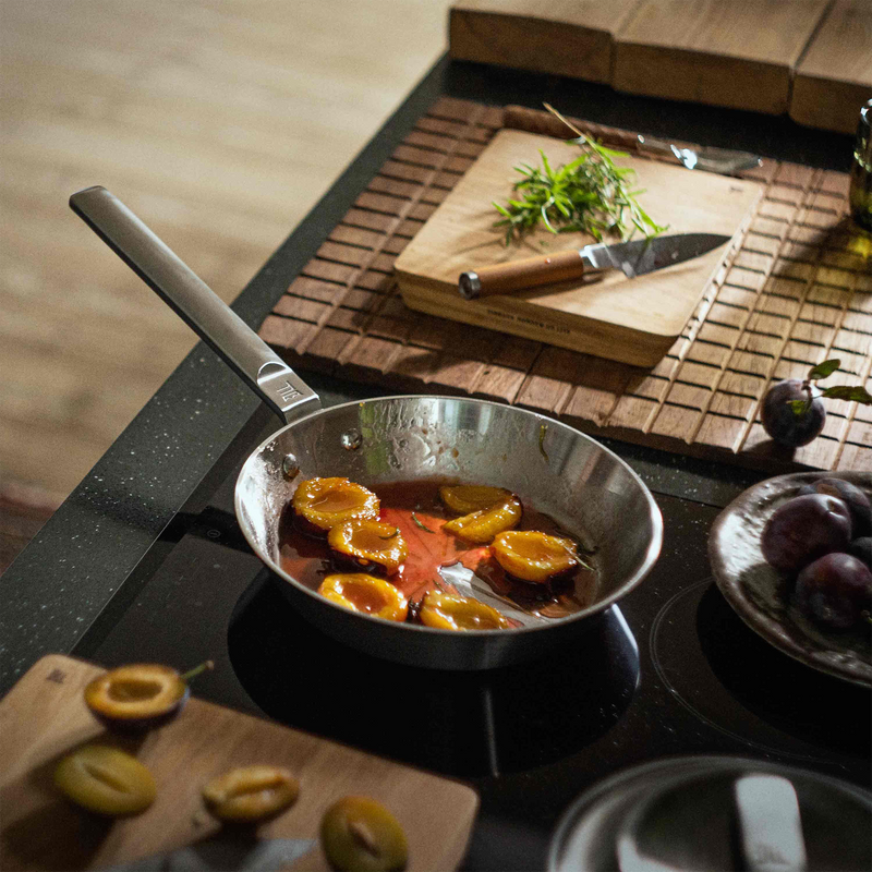 Frying pan with food on a stove, surrounded by ingredients and utensils on a wooden surface.
