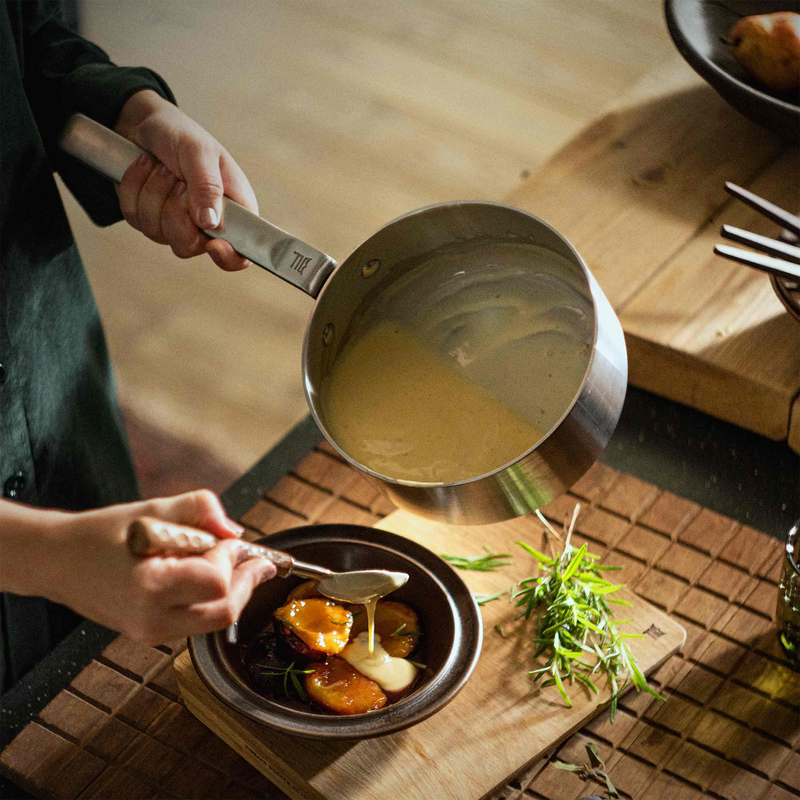 Person pouring a sauce from a small pot onto a dish on a wooden table.