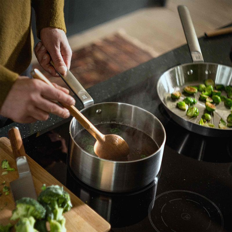 Person stirring a pot of soup on a stove with a wooden spoon, surrounded by cooking utensils and ingredients.