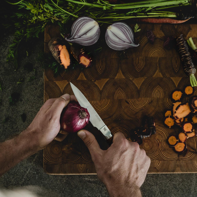 Person cutting a red onion on a wooden cutting board with various vegetables around.