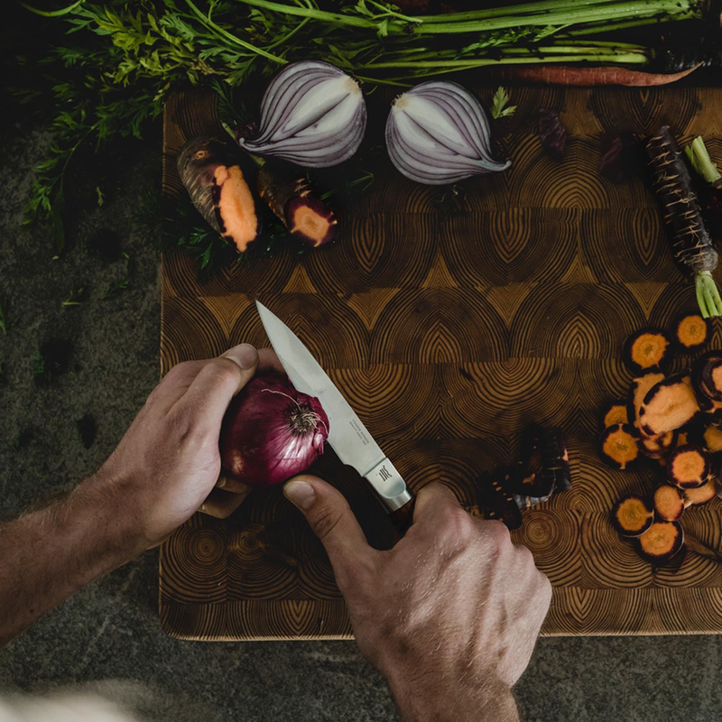 Person cutting a red onion on a wooden cutting board with various vegetables around.