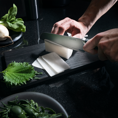 Person slicing white radish on a dark cutting board with a Shun knife, surrounded by green leaves.