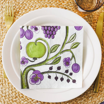 Napkin with green apple and purple grapes design on a white plate with cutlery on a woven placemat.