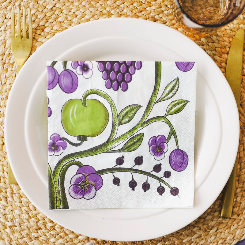 Napkin with green apple and purple grapes design on a white plate with cutlery on a woven placemat.