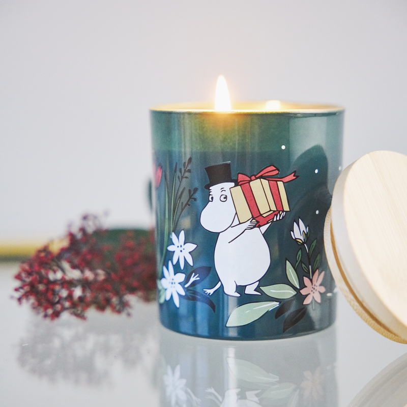 Candle in a decorative container with a character holding a gift, surrounded by flowers on a light background.
