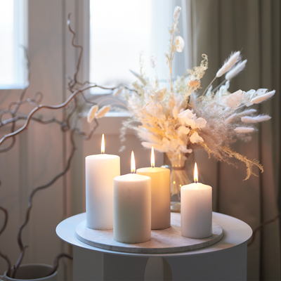 Four white candles on a small round table with a vase of dried flowers in the background.