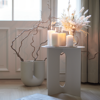 Decorative setup with candles, flowers, and branches on a white table in a bright room.