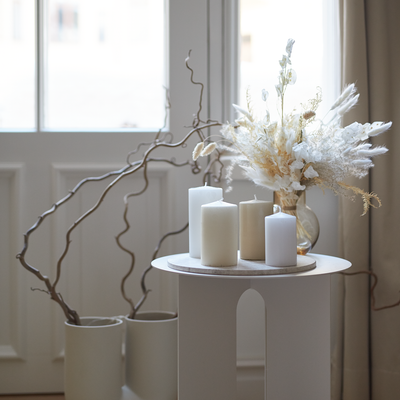 White candles and dried flowers on a small round table near a window.