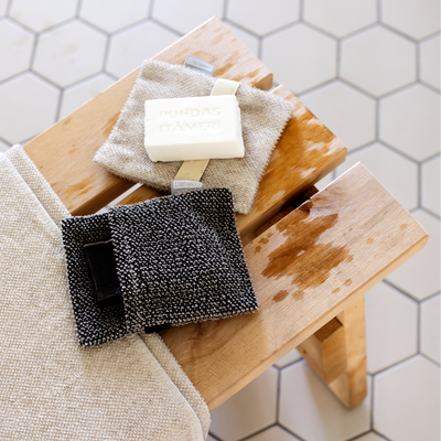 Cleaning supplies including a sponge, scrubber, and cloth on a wooden stool with a tiled bathroom background.