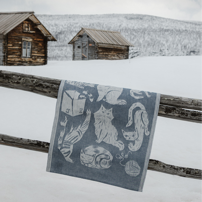 Tea towel with cat design hanging on a wooden fence in a snowy landscape with log cabins.