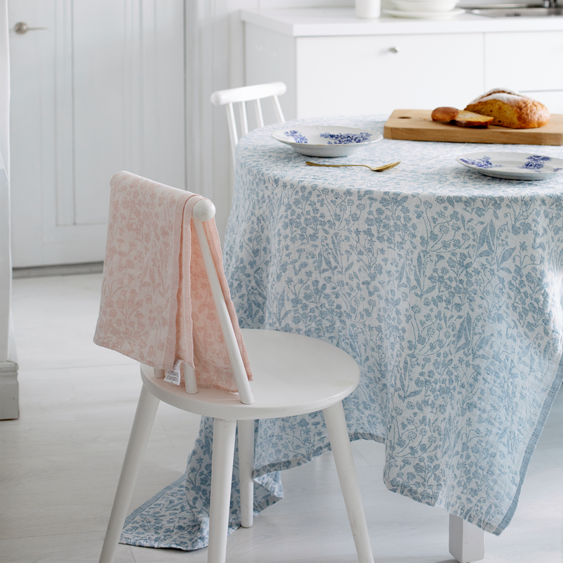 Dining room with a table covered by a floral tablecloth, a chair with a pink towel, and bread on a cutting board.