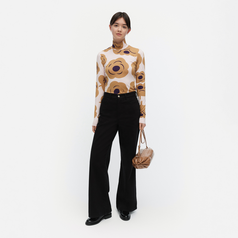 Woman wearing a patterned blouse and black pants with a beige handbag on a white background