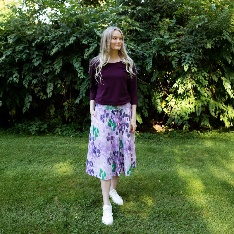 Woman in a purple top and floral skirt standing in a grassy area with greenery.