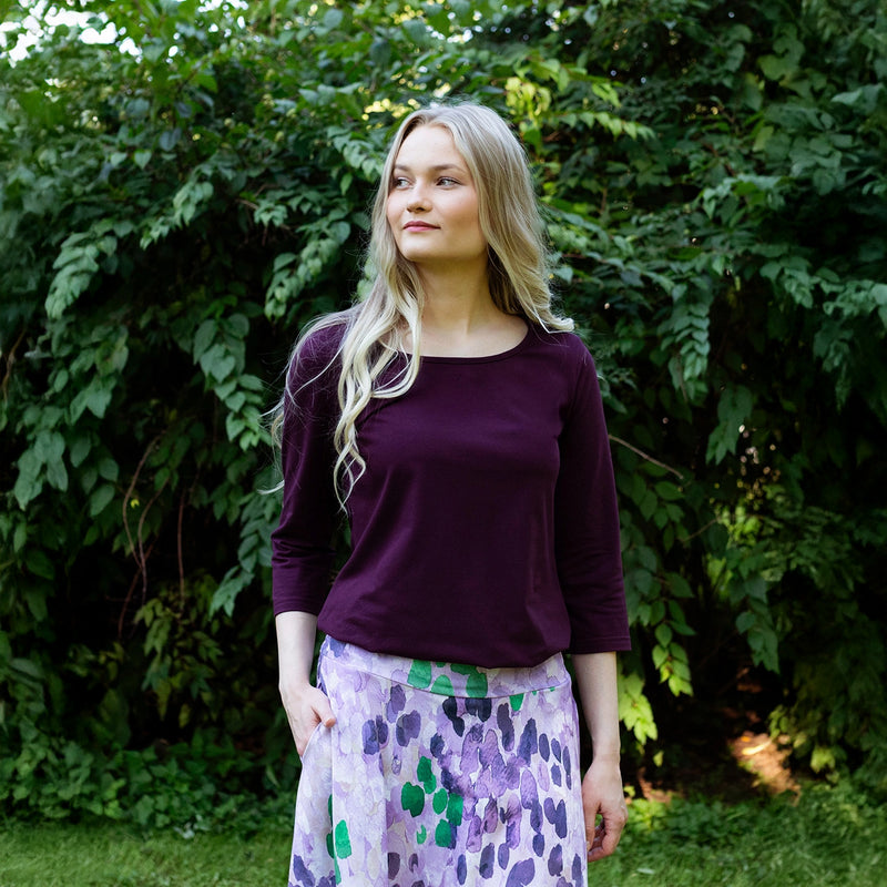 Woman wearing a purple top and patterned skirt standing in front of green foliage