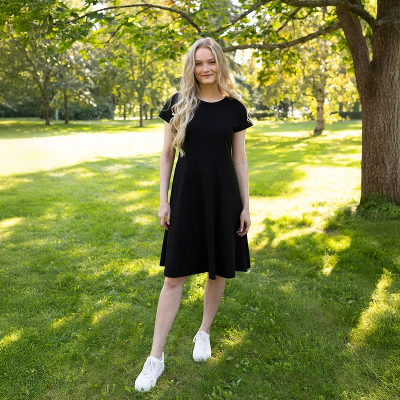 Woman in a black dress standing in a park with green grass and trees.