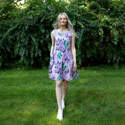 Woman in a purple floral dress standing in a grassy area with greenery.