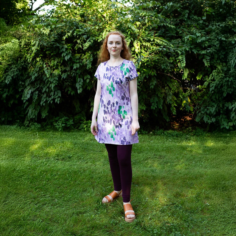 Woman wearing a purple floral dress standing in a grassy area with greenery.