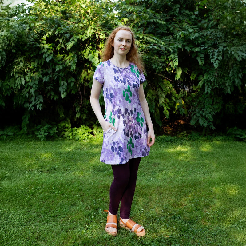Woman wearing a purple floral dress standing in a grassy area with greenery.