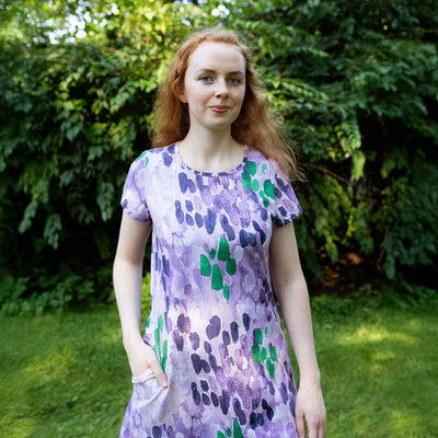 Woman wearing a purple dress with green patterns standing outdoors in a garden.