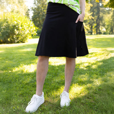 Person wearing a black skirt and white sneakers on grass with trees in the background