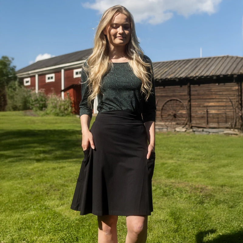 Woman in a green top and black skirt standing in a grassy area with wooden buildings in the background.