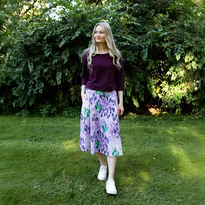 Woman in a purple top and floral skirt standing in a grassy area with greenery.