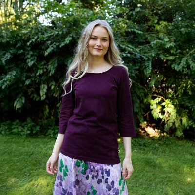 Woman wearing a purple long-sleeve top and patterned skirt standing outdoors with greenery in the background
