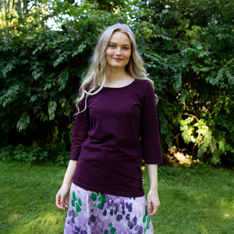 Woman wearing a purple long-sleeve top and patterned skirt standing outdoors with greenery in the background