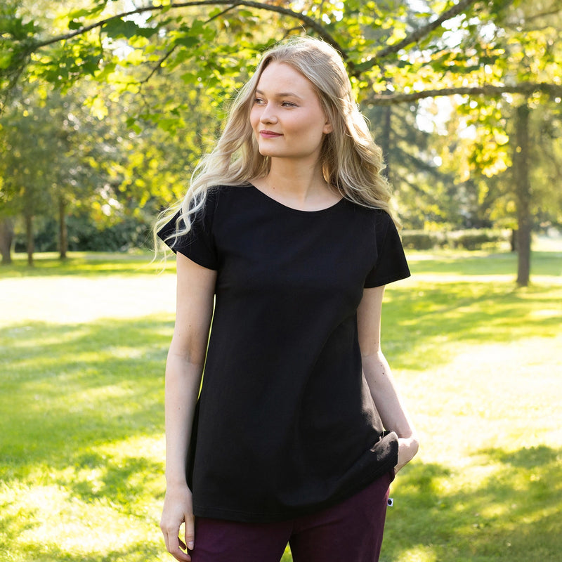 Woman wearing a black t-shirt and maroon pants standing in a park with green trees and grass.