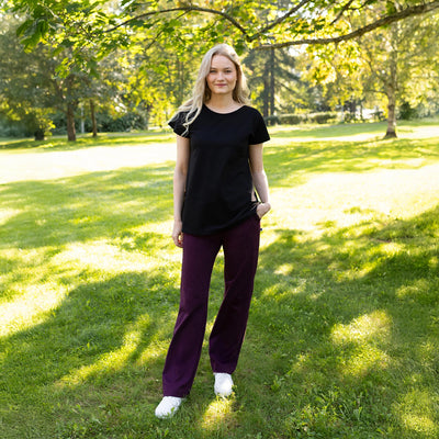 Woman in black top and purple pants standing in a park with green grass and trees.