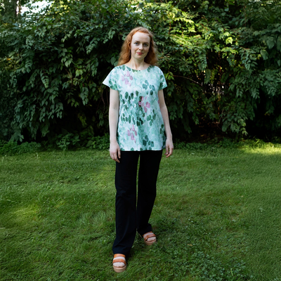Woman wearing a floral blouse and black pants standing in a grassy area with greenery.