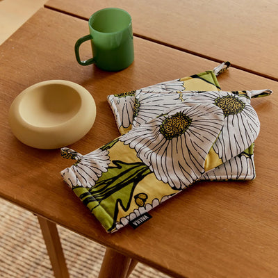 Two floral-patterned pot holders on a wooden table with a green mug and beige bowl.